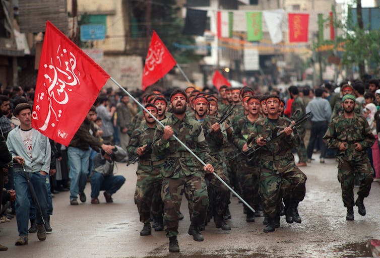 A group of men in army fatigues carrying red flags with Arabic writing on it.